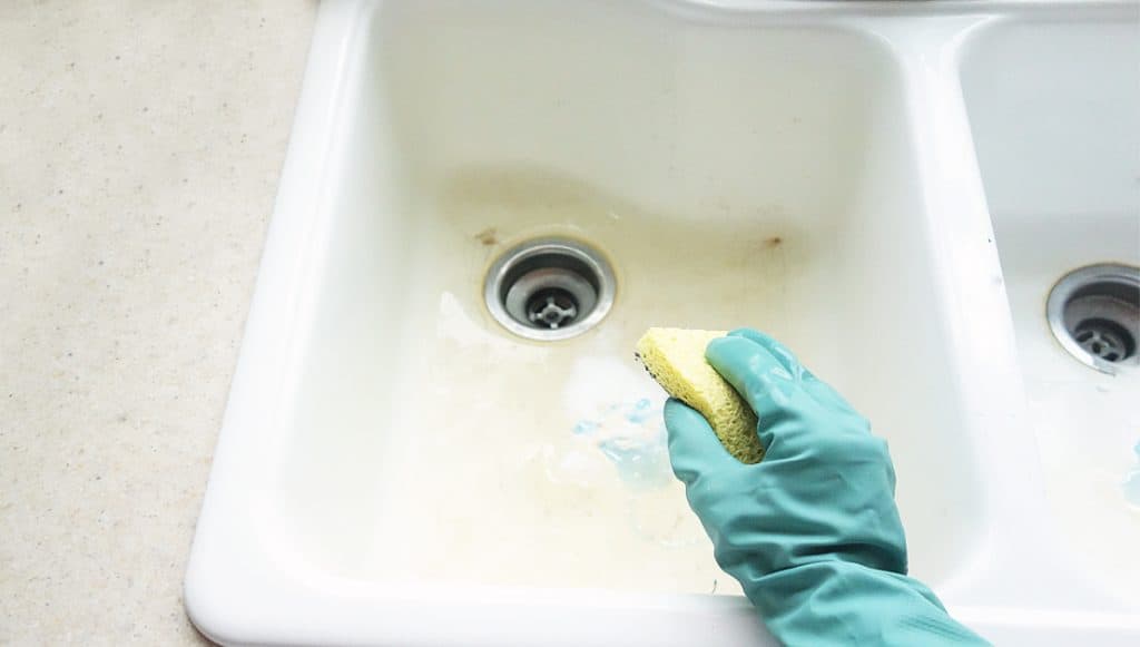 a hand with a rubber cleaning glove holds a yellow scrubby cleaning a white kitchen sink