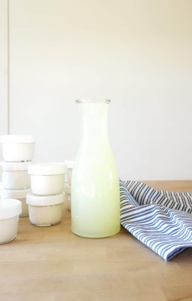 A glass bottle of whey liquid with a striped farmhouse towel and mason jars of homemade yogurt