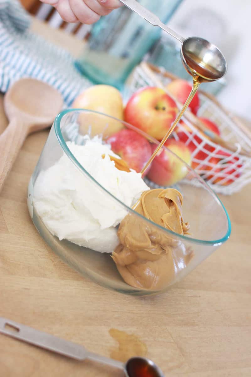 a bowl has a dollop of peanut butter and Greek yogurt. a hand is pouring in a Tablespoon of syrup. in the background is a basket of apples and a striped dish towel