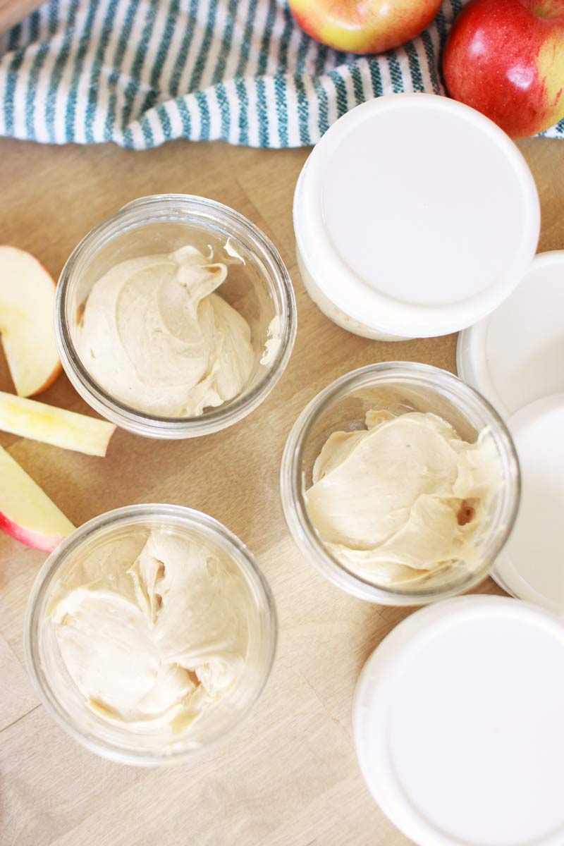individual mason jars of peanut butter yogurt dip. there are lids next to them and slices of apples