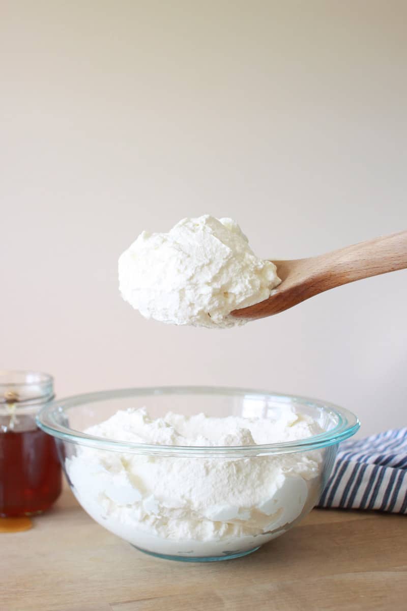 a wooden spoon with a large dollop of whipped cream and a bowl of whipped cream. A jar of honey and a striped dish towel are behind it.