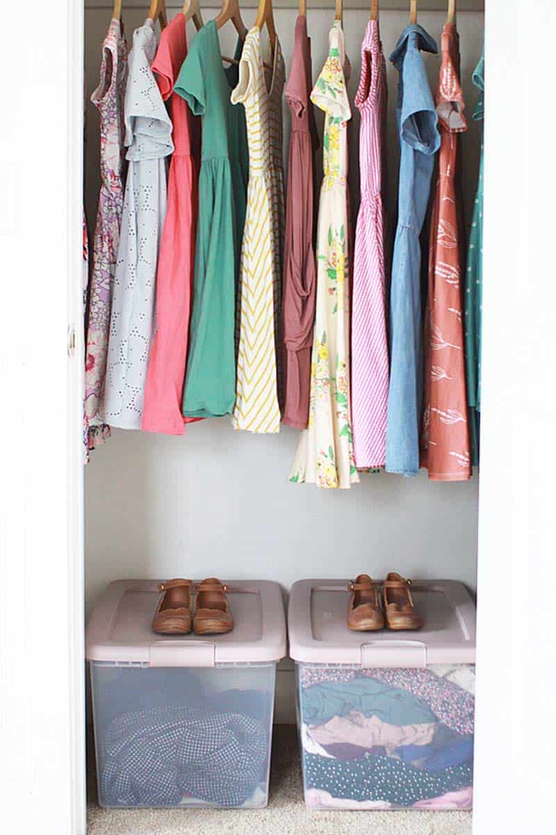 a closet with girls dresses hanging on hangers and bins on the floor with church shoes laid on top of them