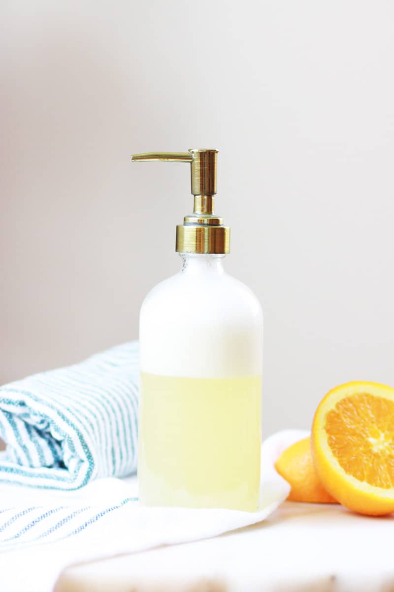 a glass bottle of homemade orange face soap with a cut orange and striped towel in the backgroung