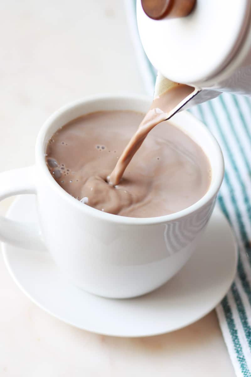 homemade cocoa being poured out of a cocoa pot into a white hot cocoa cup