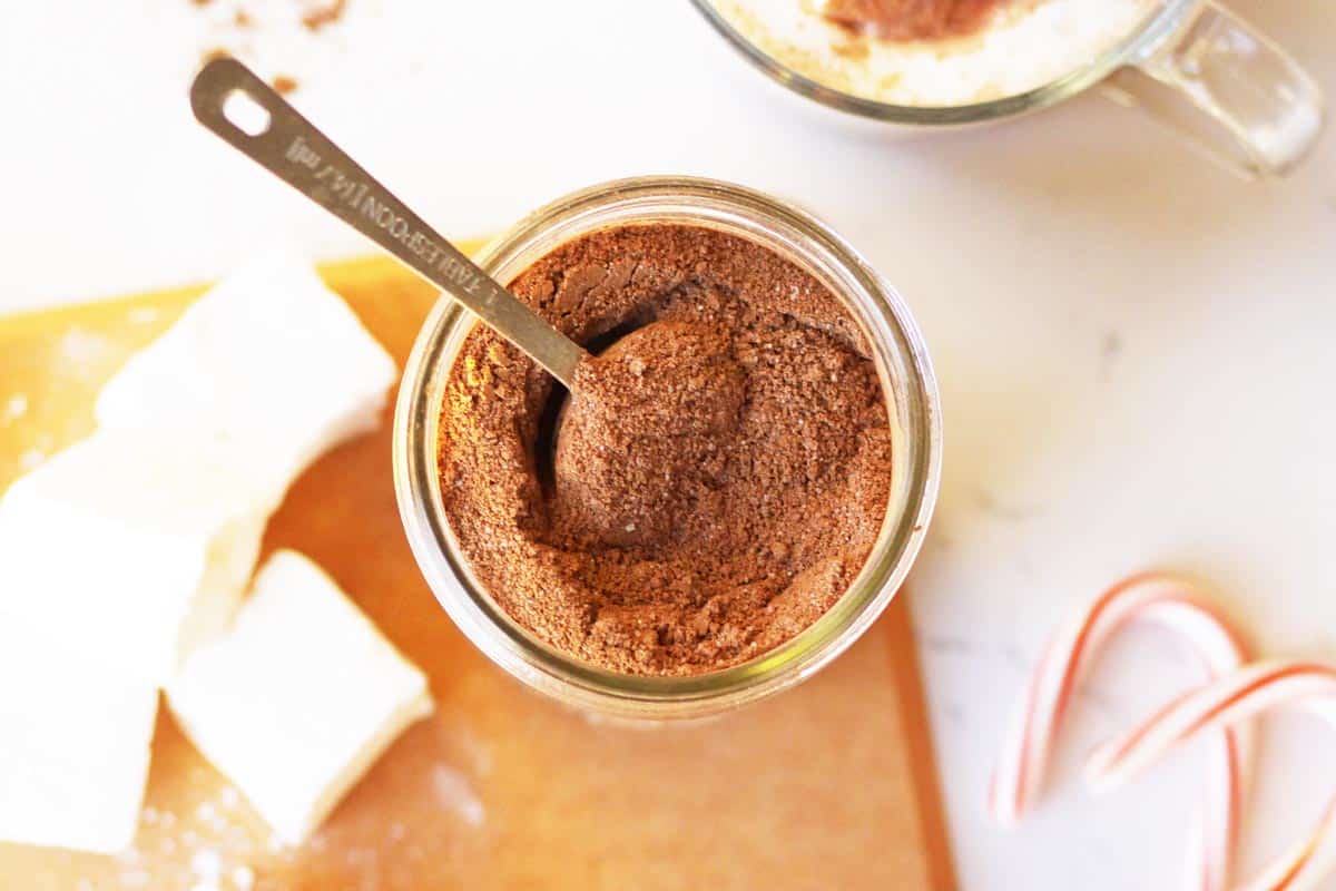 a mason jar of hot cocoa mix with cocoa powder, a cup of hot chocolate, candy canes and marshmallows next to it