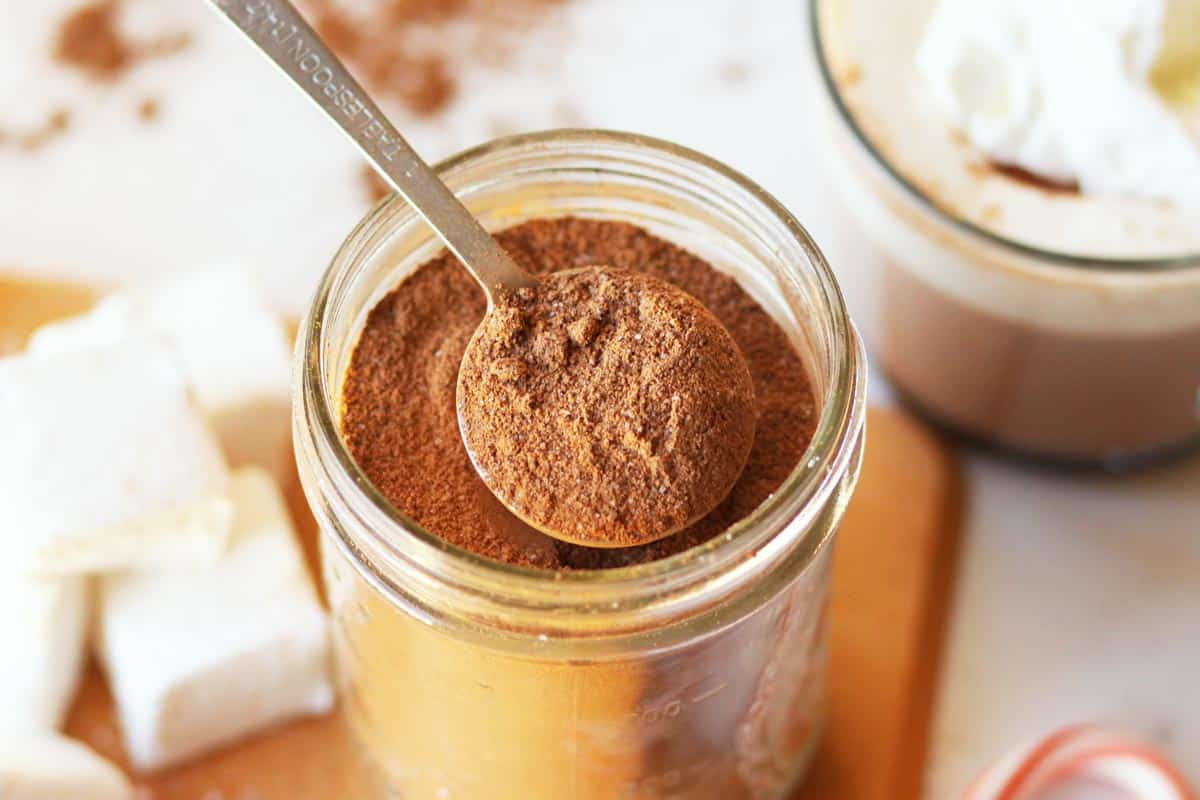 a mason jar of hot cocoa mix with cocoa powder, a cup of hot chocolate, candy canes and marshmallows next to it