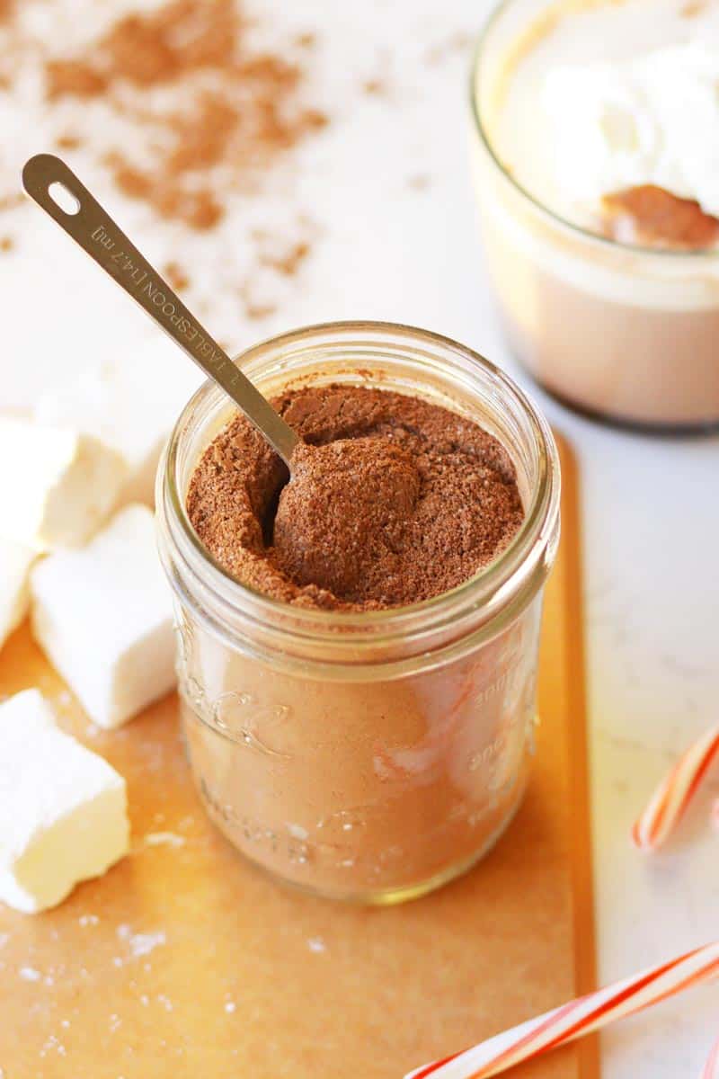 a mason jar of hot cocoa mix with cocoa powder, a cup of hot chocolate, candy canes and marshmallows next to it