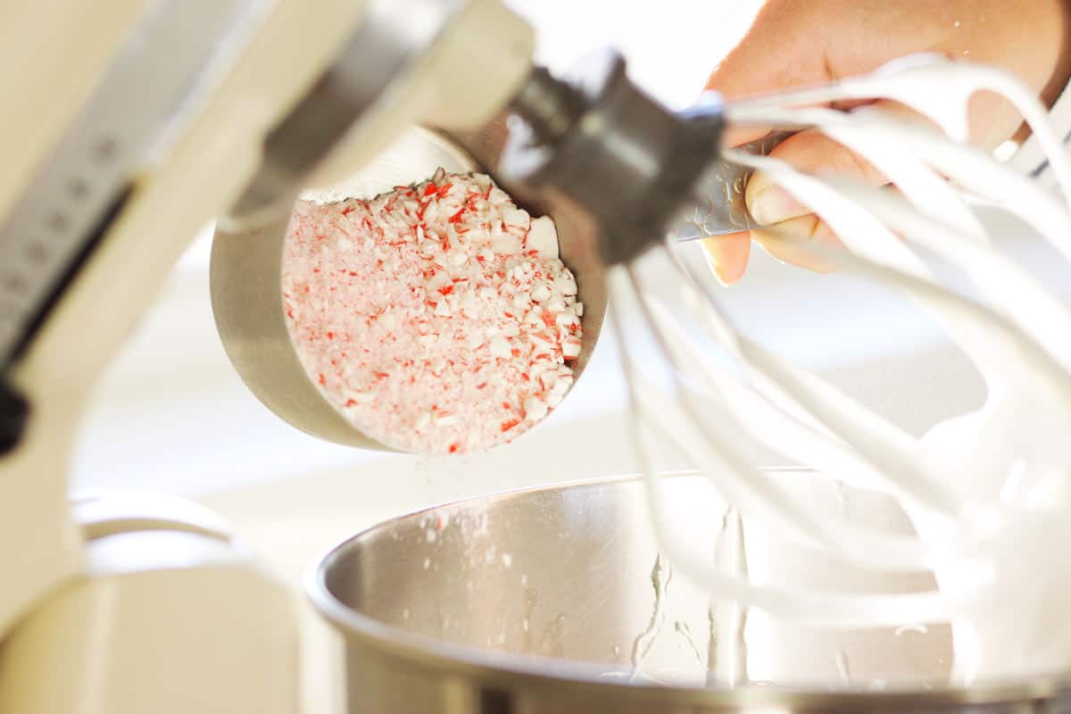 crushed candy canes being poured into a kitchen aid mixer bowl with homemade marshmallow fluff in it