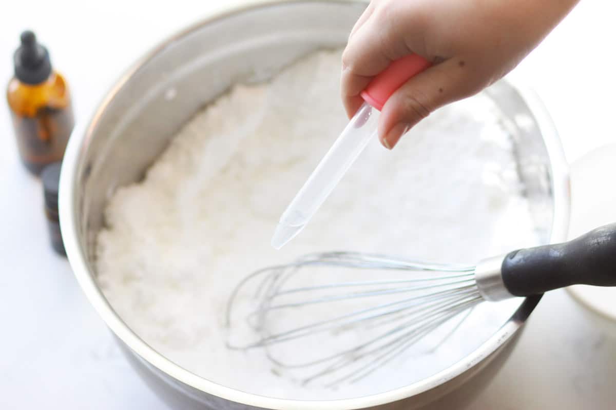 a hand is holding an eye dropper filled with vinegar over a bowl of baking soda mixed with citric acid. An essential oil bottle is nearby