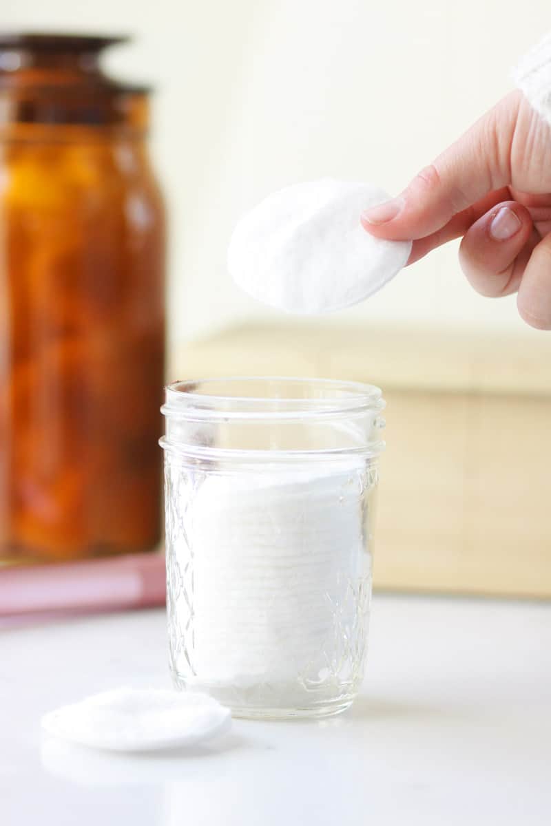 a jelly jar with a stack of homemade makeup remover wipes inside. A hand is holding one round makeup wipe.