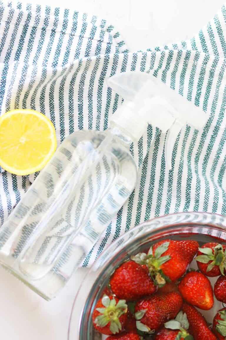 a glass spray bottle on a striped towel and a bowl of strawberries in a bowl of water and vinegar