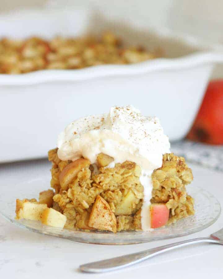 a slice of apple baked oatmeal on a plate with a casserole dish in the background. The slice is topped with whipped cream and cinnamon.