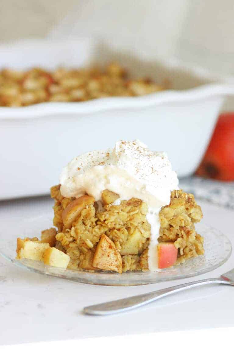a slice of apple baked oatmeal on a plate with a casserole dish in the background. The slice is topped with whipped cream and cinnamon.