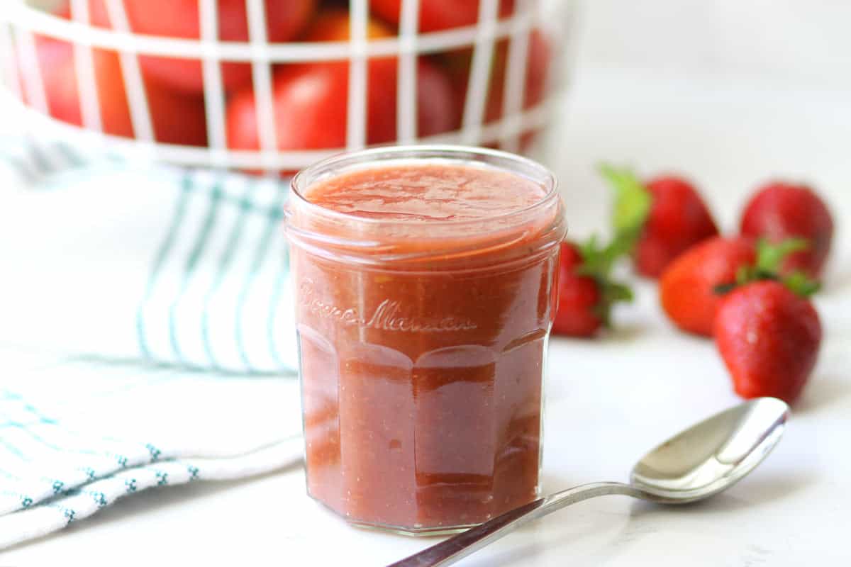 a jar of strawberry applesauce with a basket of apples and strawberries laying around