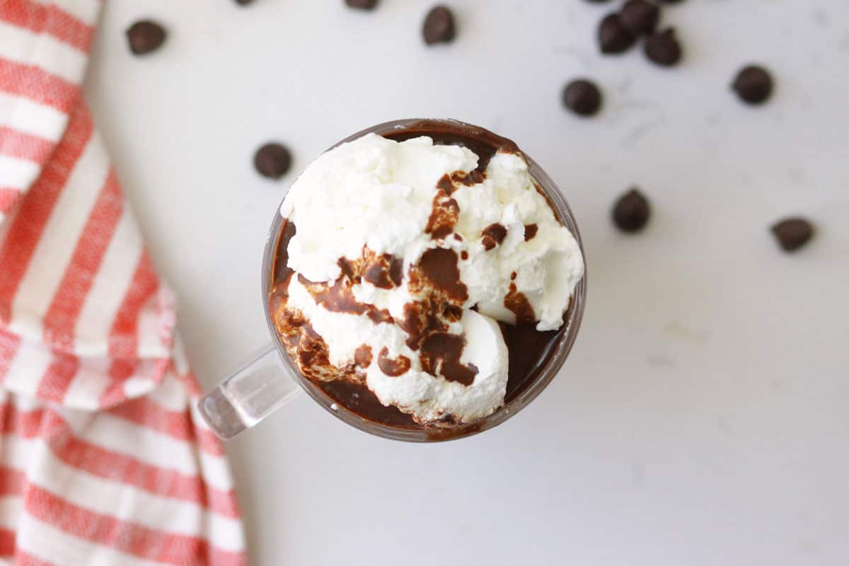 a cup of creamy hot chocolate with a red and white striped towel behind it, chocolate chps scattered around and whipped cream on top