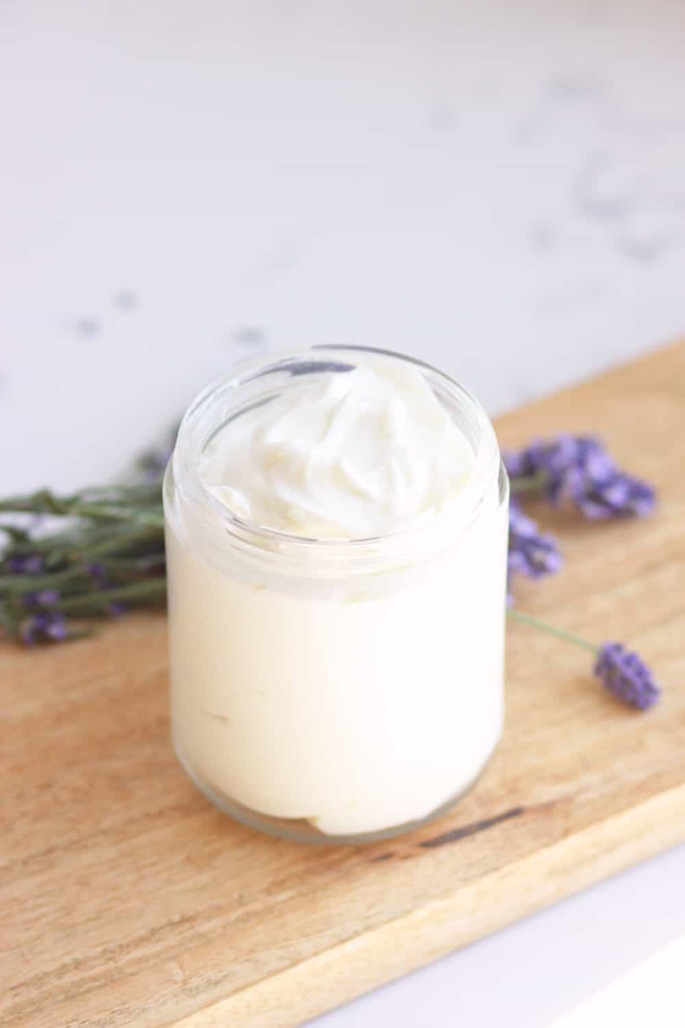 a glass jar of lotion with lavender buds laying around it
