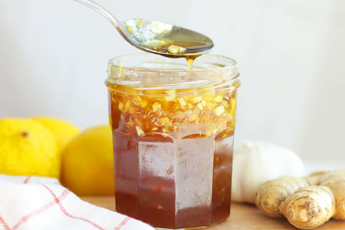 a jar of homemade cough syrup with honey, minced garlic and ginger in it. A lemon and garlic are on a cutting board behind it.