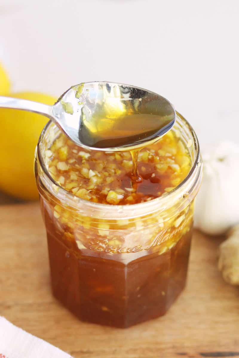 a jar of homemade cough syrup with honey, minced garlic and ginger in it. A lemon and garlic are on a cutting board behind it.