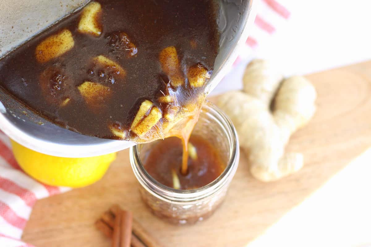 ginger, honey, cinnamon and olive oil being poured from a saucepan into a jar. Ginger root and cinnamon sticks in the background