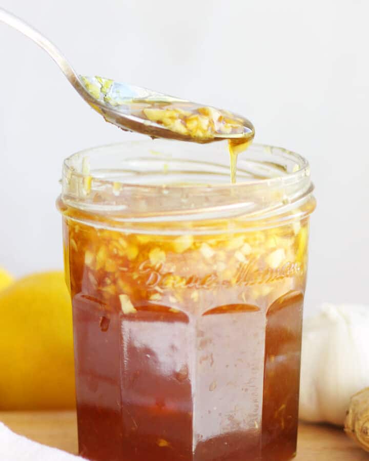 a jar of homemade cough syrup with honey, minced garlic and ginger in it. A lemon and garlic are on a cutting board behind it.