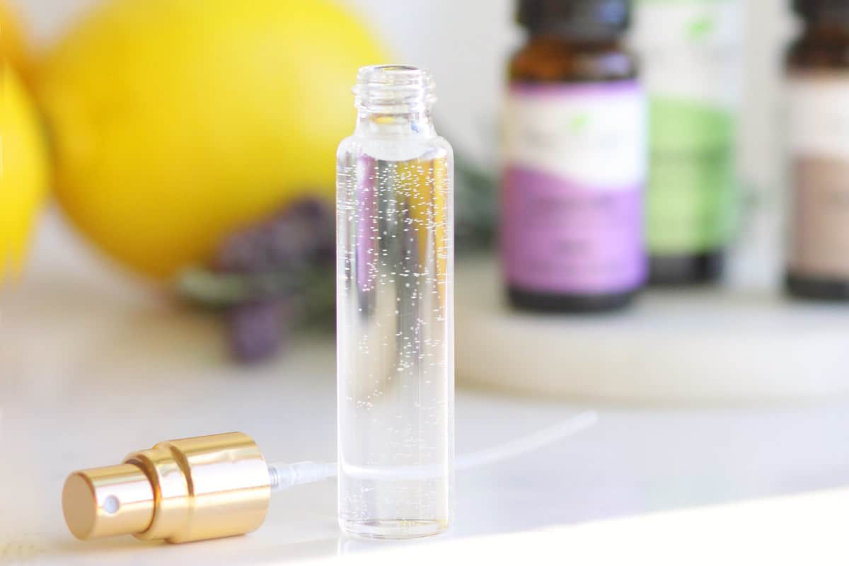 a clear spray perfume bottle is set on a table with fresh lavender, rosemary, essential oil bottles and lemons next to it.