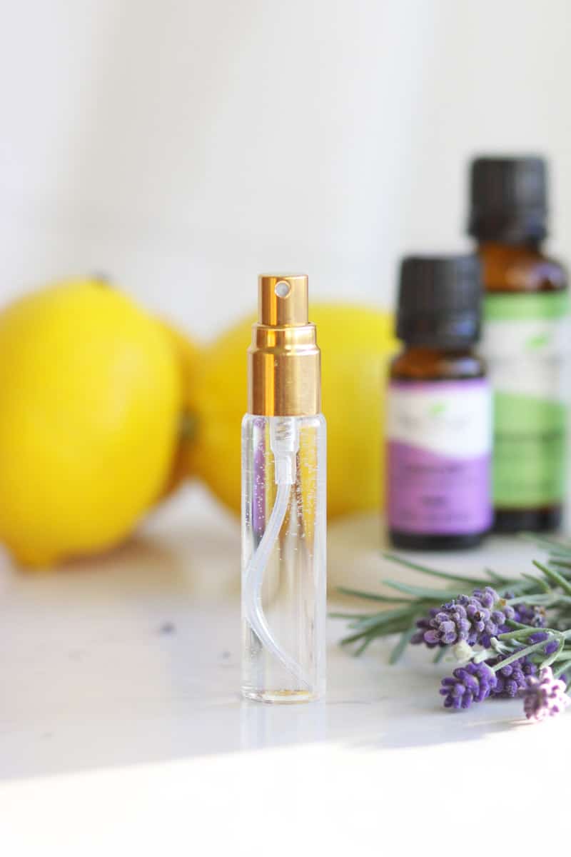 a clear spray perfume bottle is set on a table with fresh lavender, rosemary, essential oil bottles and lemons next to it.