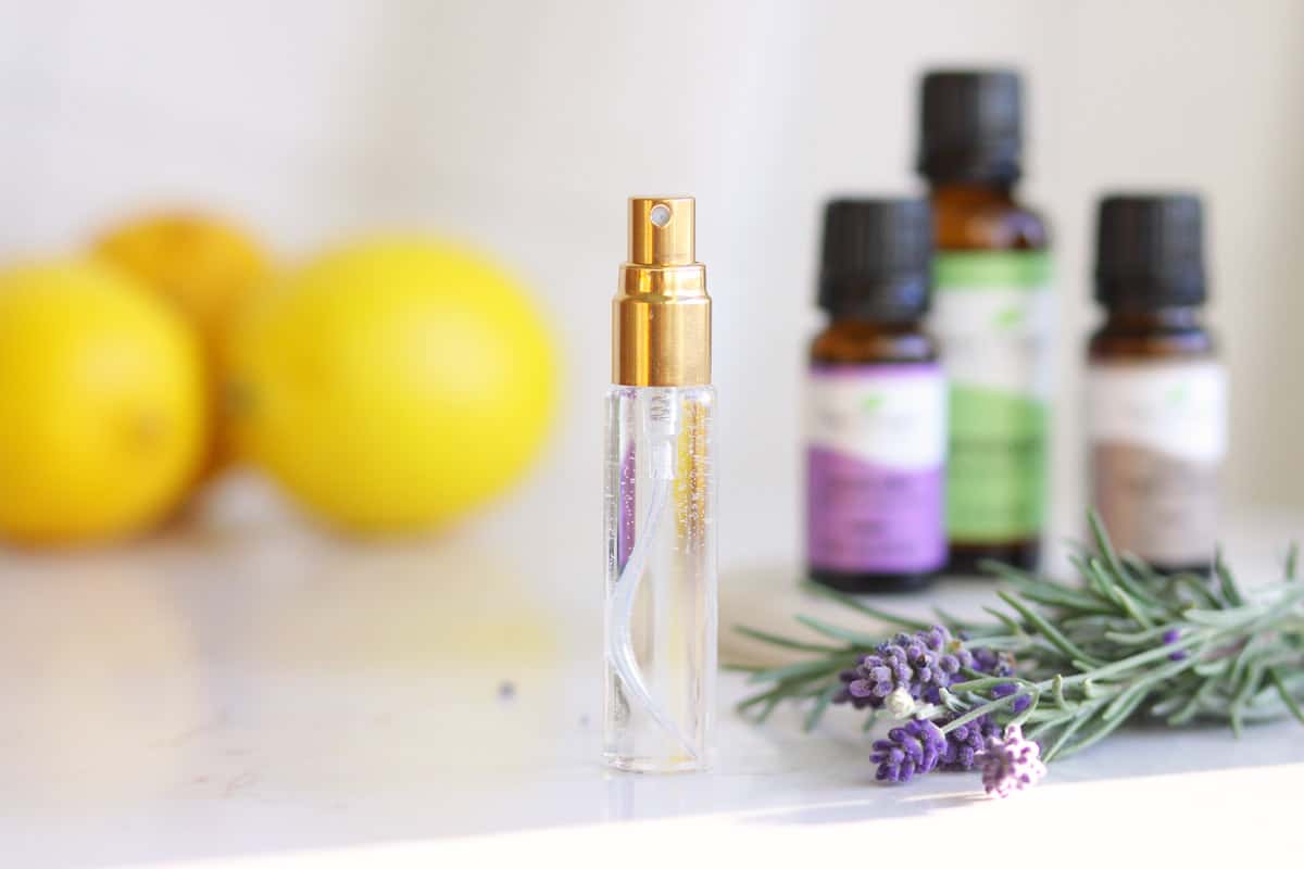 a clear spray perfume bottle is set on a table with fresh lavender, rosemary, essential oil bottles and lemons next to it.