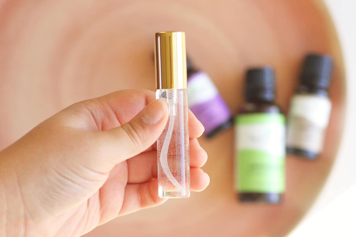 a hand holding a clear perfume bottle over a pink plate with three essential oil bottles on it.