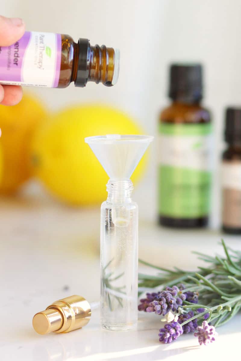 essential oils being dripped into a small spray perfume bottle. Fresh lavender, rosemary, and lemons are on the table next to it.