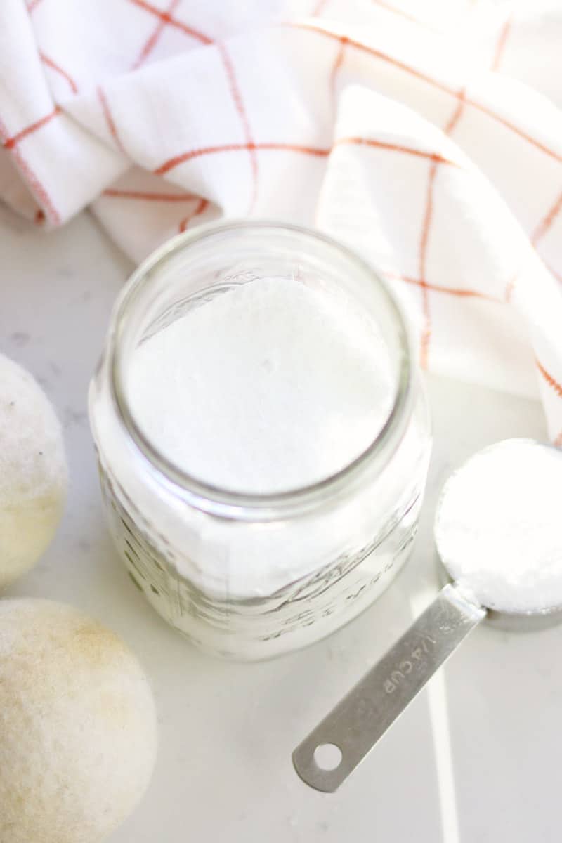 a jar of homemade scent booster made with baking soda and salt. Wool balls and clean towels are laying next to it as well as a cup measure.