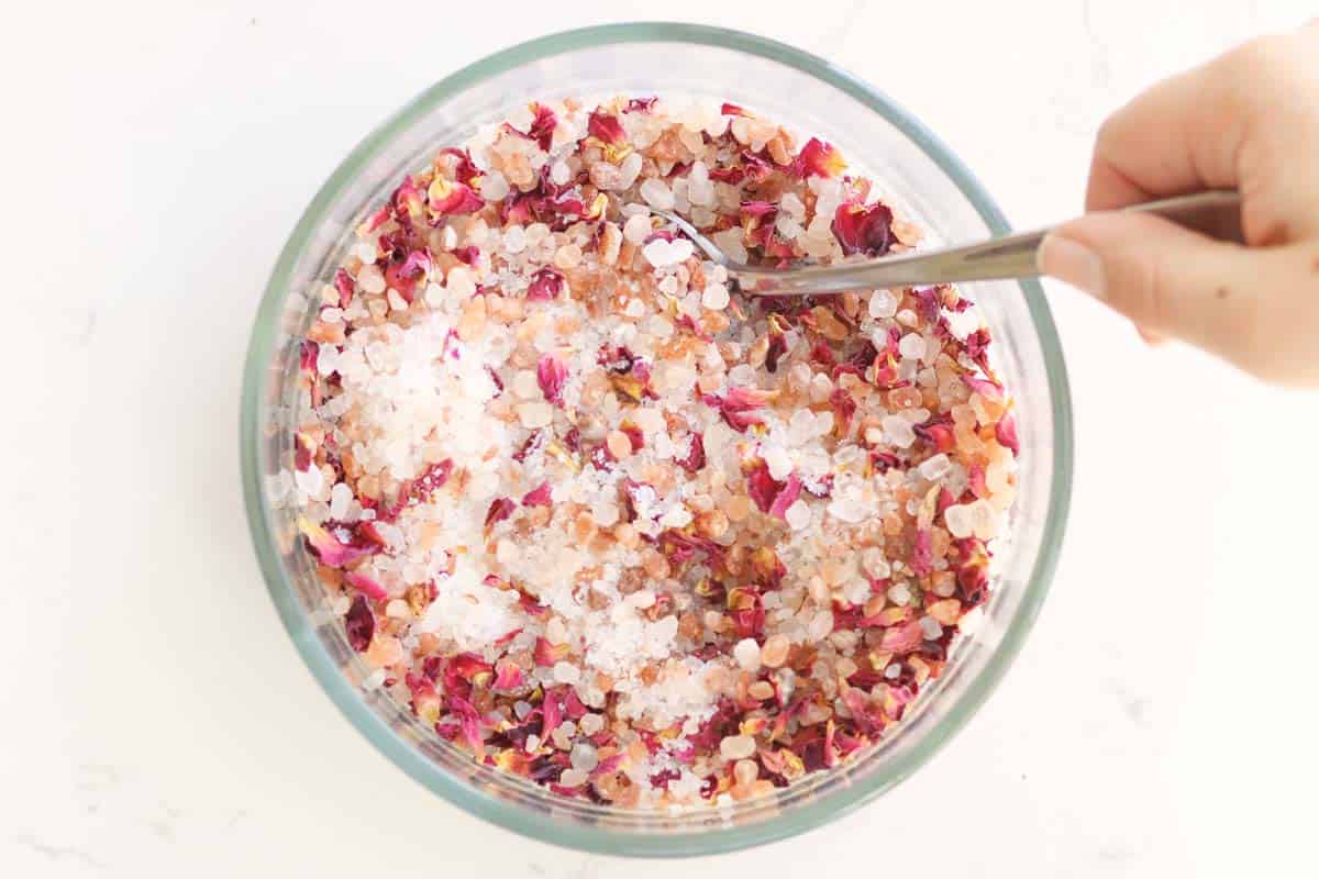 a bowl of sea salt, Himalayan pink salt and rose petals being stirred with a spoon.