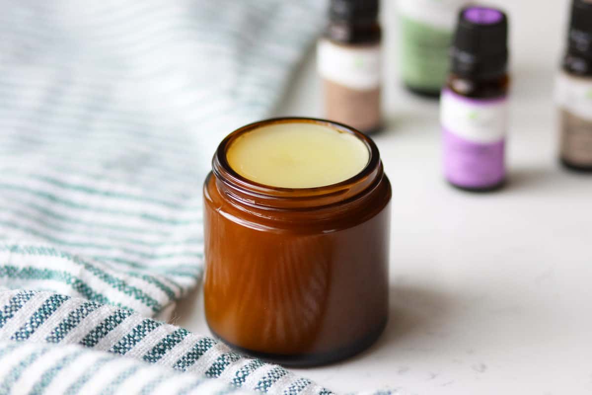 an amber glass jar with homemade vapor rub in it with a striped green and white towel laid under it and essential oils laying next to it