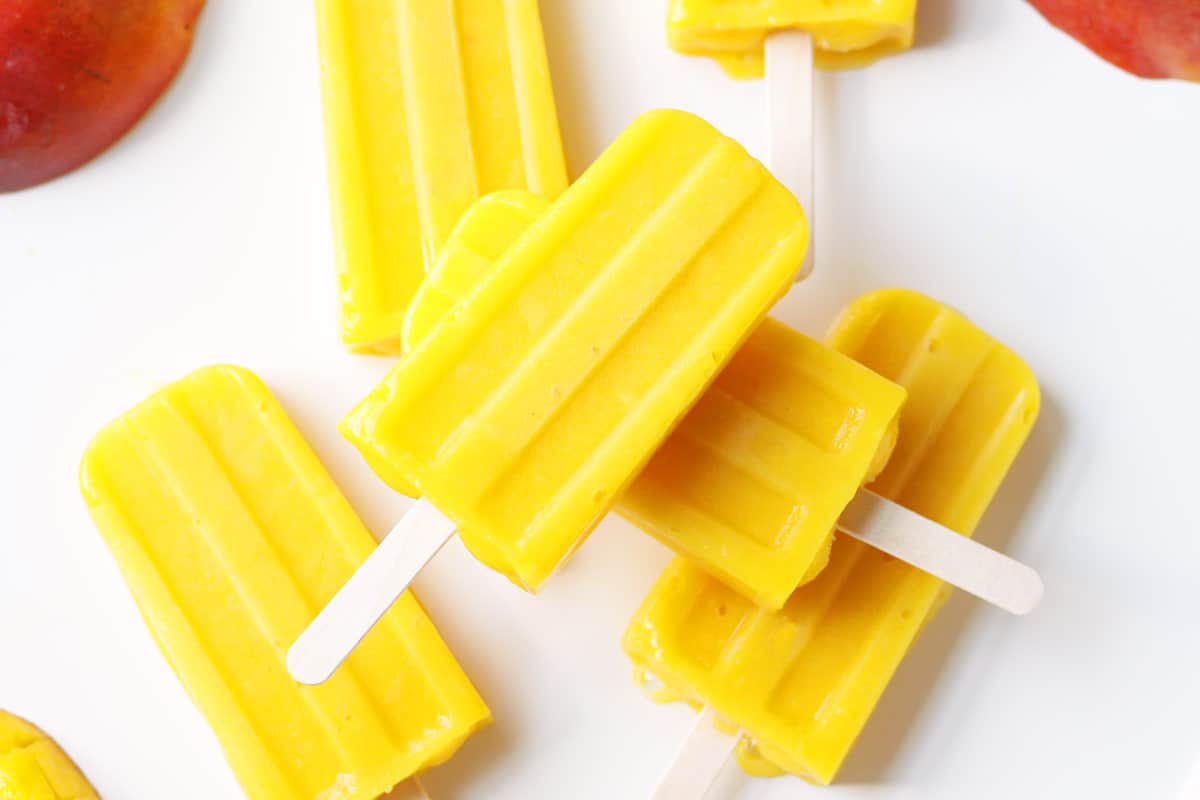 a pile of mango popsicles on a white tray with a sliced fresh mango behind them