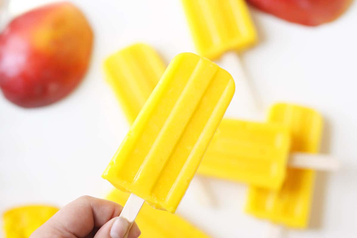 a pile of mango popsicles on a white tray with a sliced fresh mango behind them
