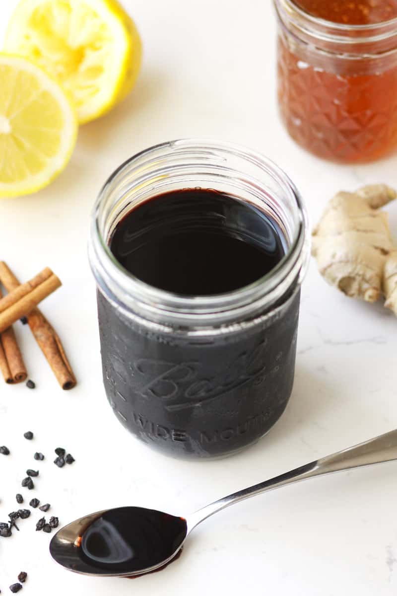 a mason jar of elderberry cough syrup with ginger, cinnamon, elderberries, honey and lemon laying next to it