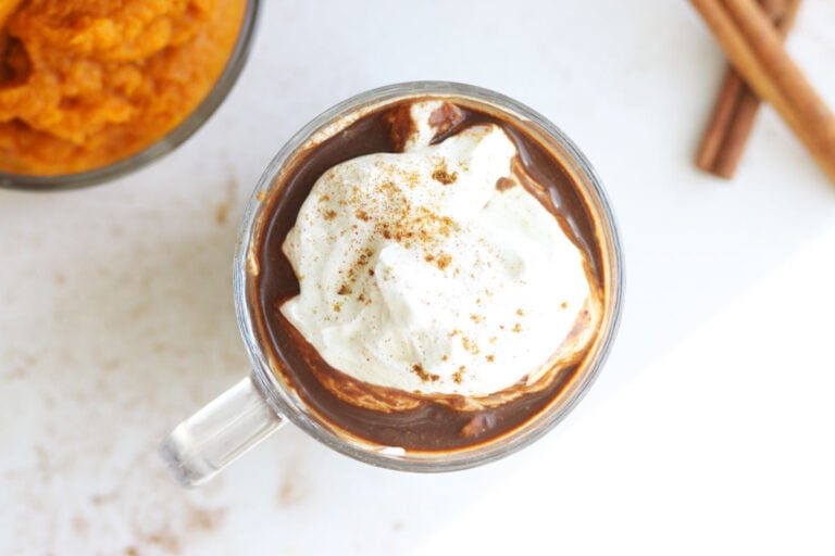 a glass mug with pumpkin spice cocoa topped with whipped cream and pumpkin pie spice sprinkled on top. A cup of pumpkin puree and cinnamon stick are laying on the table.