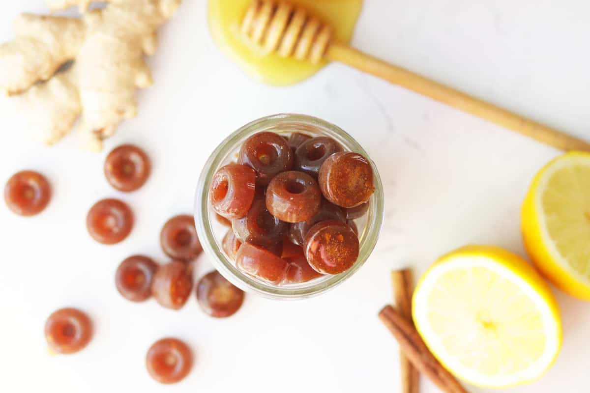 a mason jar of homemade cough drops with ginger root, honey on a honey stir stick, lemon and cinnamon sticks on the table next to it.