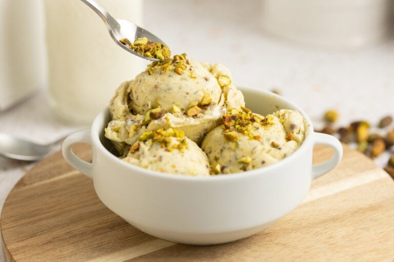 A white bowl of pistachio ice cream with a spoon in it and chopped pistachios laying around it