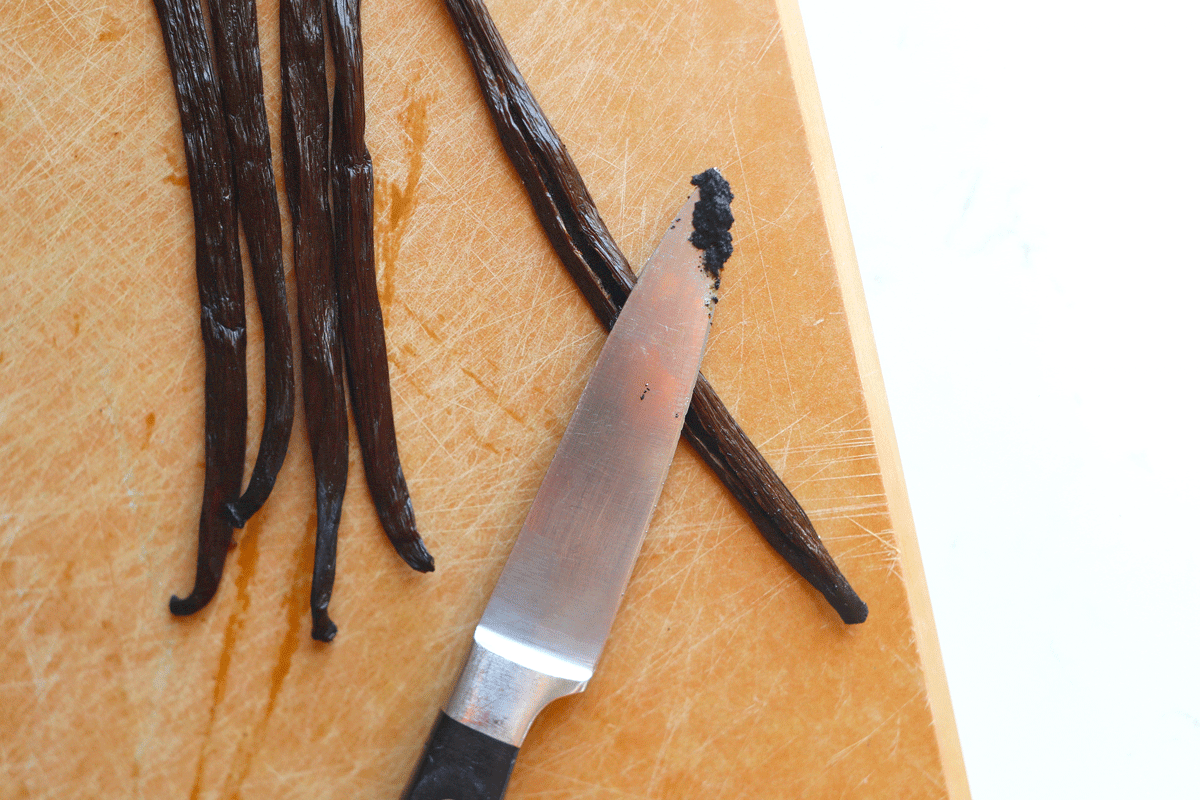 A knife with vanilla seeds on it. Whole vanilla beans are on the cutting board next to it