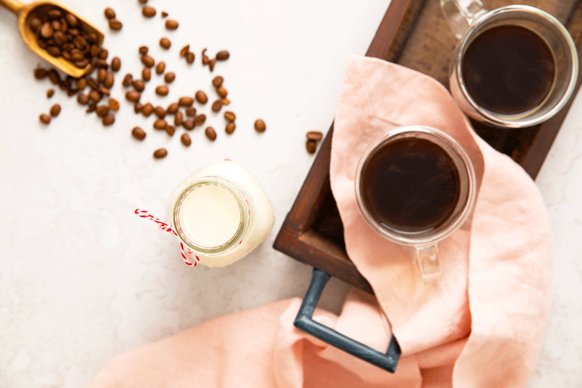 A glass bottle of homemade coffee creamer with a cup of hot coffee and coffee beans next to it.