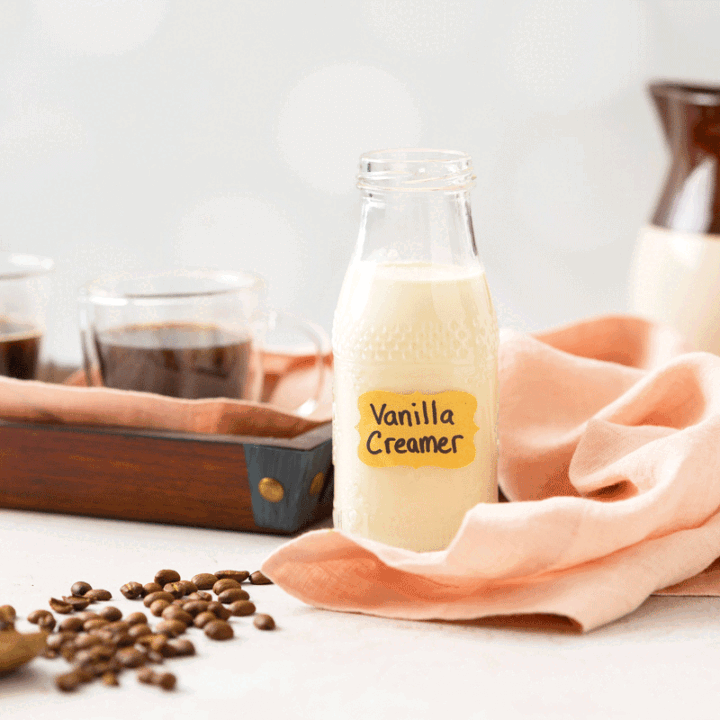 A glass bottle of homemade coffee creamer with a cup of hot coffee and coffee beans next to it.